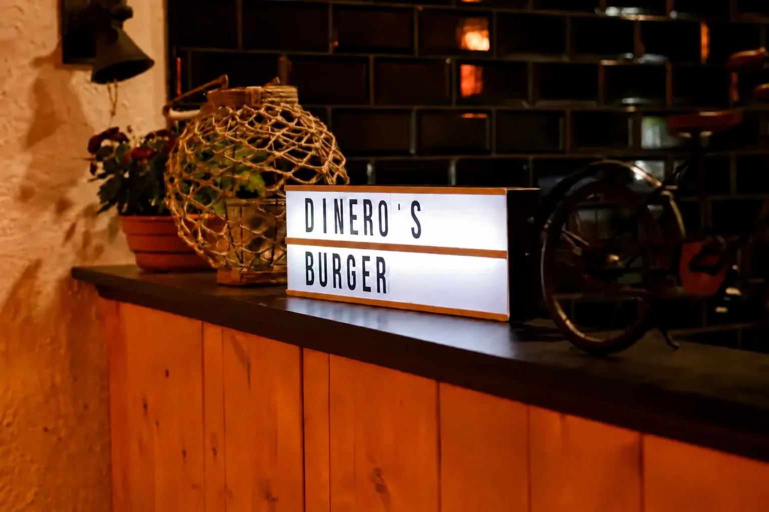 Beleuchtetes Schild mit der Aufschrift Dinero's Burger auf der Theke, daneben eine Korbflasche und eine Topfpflanze